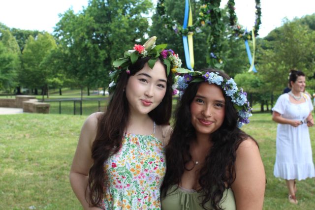 Two women wear a crown of flowers in front of a maypole. 