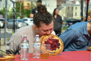 A participate eats a strawberry pie in the Philly AIDS Thrift's annual pie eating contest. They are biting their teeth into the crust. They are holding the pie up with their teeth, so it's standing up and facing forward.